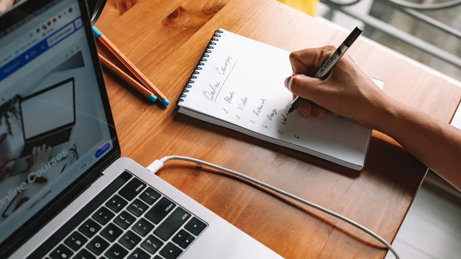 Pessoa sentada, escrevendo anotações sobre cursos online num caderno, com um laptop aberto ao lado