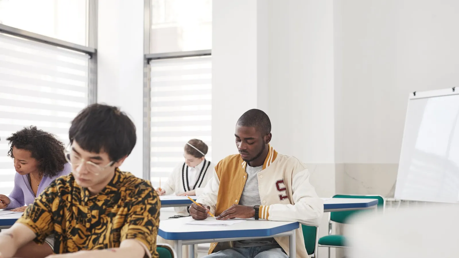 Estudantes sentados em carteiras individuais, concentrados, fazendo uma prova numa sala clara